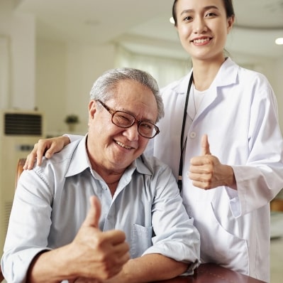 Smiling elderly man giving a thumbs up while sitting next to a female doctor who is also giving a thumbs up.